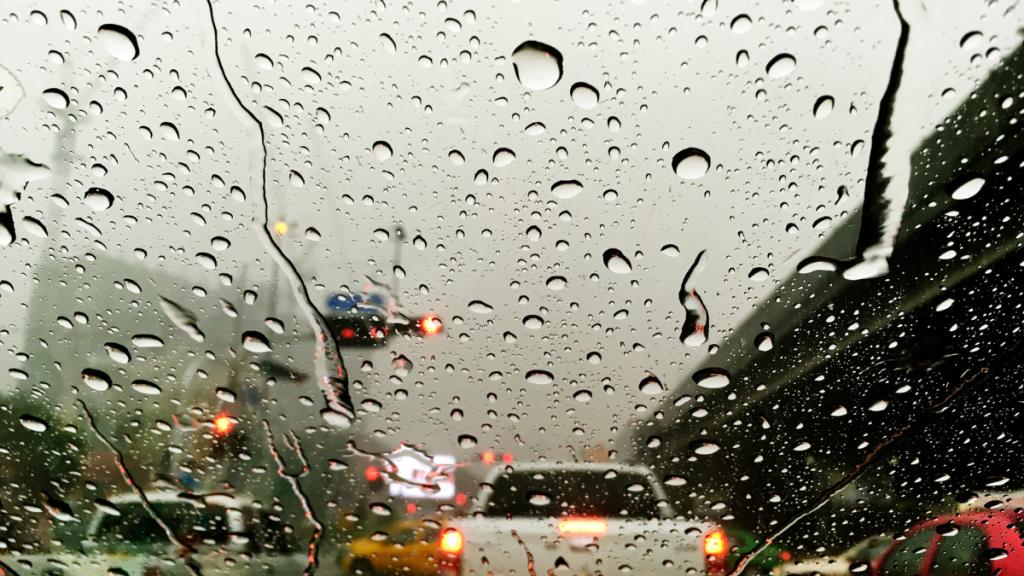 Previsão do Tempo no estado de São Paulo: vista de carros e prédios através de uma janela com gotas de chuva, indicando tempo chuvoso e mudanças nas condições ao longo da semana, com frio nas manhãs.