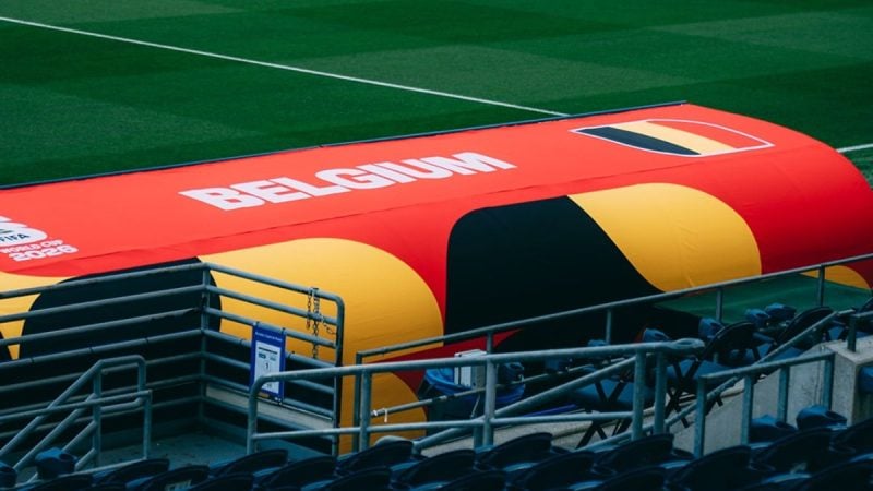 Bandeira vermelha e amarela da FIFA em um estádio, com assentos e arquibancada ao fundo, ilustrando o debate sobre a liberação de Balogun pela FIFA e críticas da federação belga.