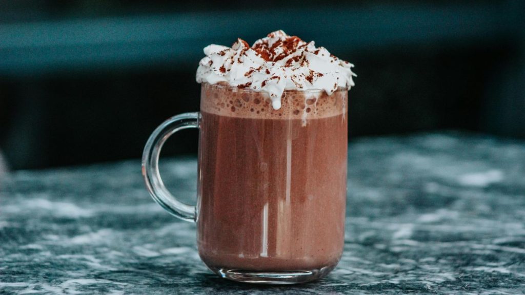 Dia do Chocolate: chocolate quente cremoso em copo de vidro com cobertura de chantilly e raspas de chocolate, servido em mesa de inverno, com fundo escuro ao fundo.
