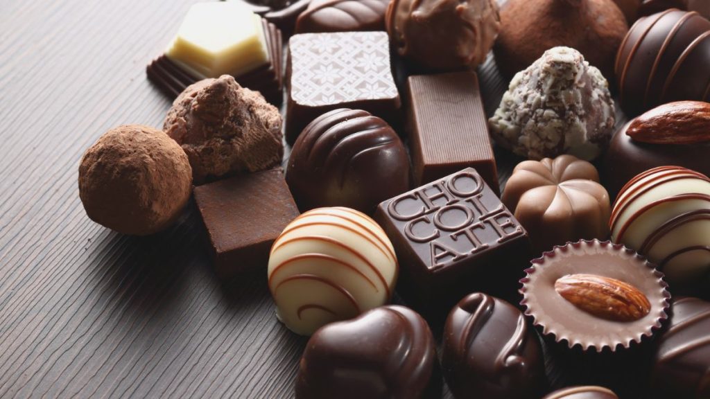 Dia do Chocolate com bombons e chocolates variados sobre fundo de madeira claro, incluindo trufas, barras e doces ao redor de confeitos decorados.