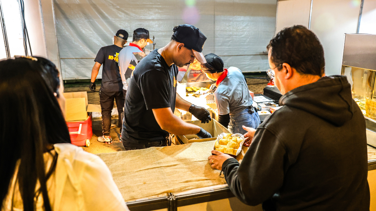 Pessoas servindo e degustando comidas em uma barraca durante o Festival Gastronômico de Inverno, em Santa Bárbara, com fila e preparação de alimentos ao ar livre
