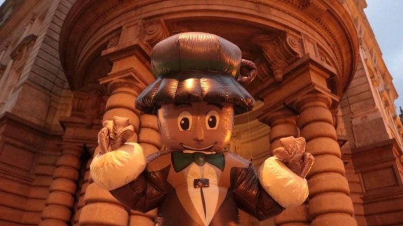 Mascote de terno e gravata, com chapéu de café e bigode de espuma, segurando um grande xícara inflável em cenário cenográfico com arcos e fachada de pedra, ideal para divulgação do Festival Santos Café com gastronomia, lazer e música no feriado.