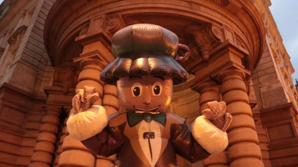 Mascote de terno e gravata, com chapéu de café e bigode de espuma, segurando um grande xícara inflável em cenário cenográfico com arcos e fachada de pedra, ideal para divulgação do Festival Santos Café com gastronomia, lazer e música no feriado.