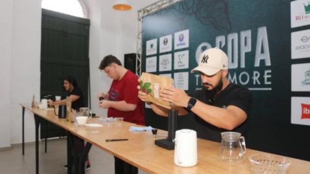 Participantes preparando café durante o Festival Santos Café, com pessoas ao lado de uma mesa de baristas, equipamentos e sacos de grãos sendo manuseados em um evento.