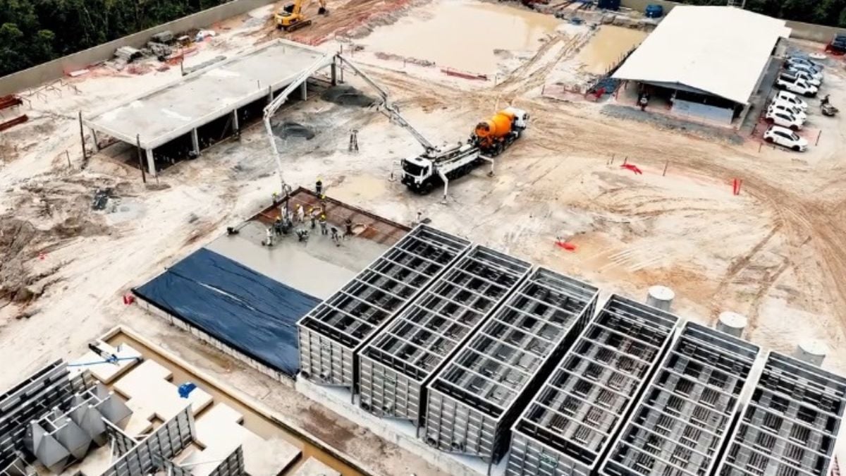 Vista aérea de obra industrial com estrutura metálica e grandes caixas de tratamento de água, máquinas e caminhões ao fundo, ilustrando investimentos da Sabesp na Baixada Santista para maior segurança hídrica.