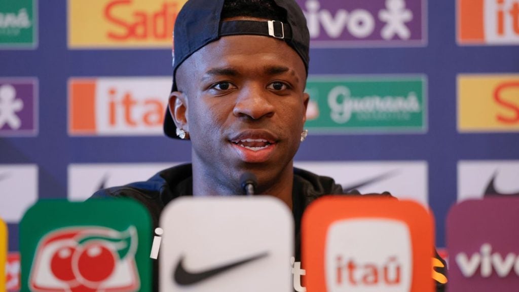Vinícius Júnior em coletiva de imprensa da Seleção Brasileira, sorrindo diante de logotipos da CBF e do patrocínio da Nike e do Itaú ao fundo, reforçando a ideia de brasil vai voltar para copa com foco no ciclo até 2030.