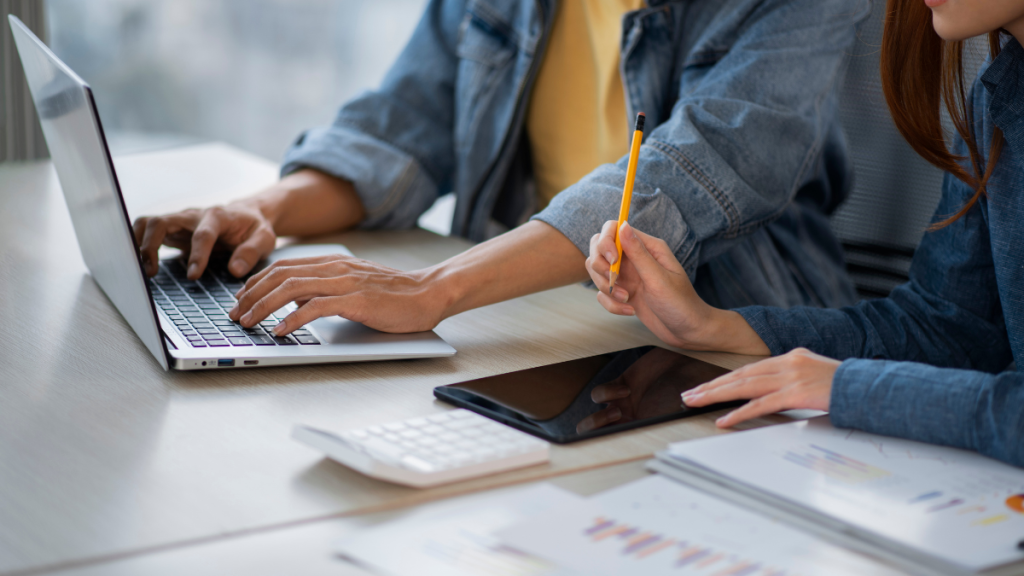 Estudantes em mesa de trabalho com notebook e documentos, enquanto uma jovem escreve em um caderno e outra digita no computador, sugerindo oportunidades de estágio TRE-SP para alunos do ensino médio.