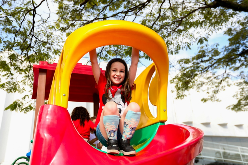 Criança sorrindo em um brinquedo de playground colorido ao ar livre, com ambiente escolar ao fundo, em divulgação do Super Férias Sesi com inscrições para Santos, Cubatão e Registro.