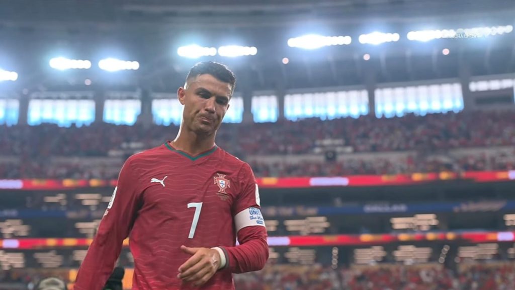 Cristiano Ronaldo em campo, usando a camisa vermelha com o número 7, durante uma partida em estádio com arquibancadas e refletores ao fundo.