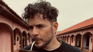 Daniel de Oliveira em foto ao ar livre, com cabelo cacheado e barba, usando camiseta escura, em frente a um conjunto de arcos e construções com tons terrosos.