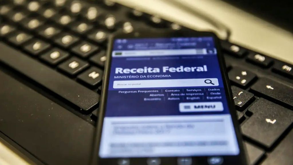 A imagem mostra um celular sobre um teclado de computador com a tela do sistema “Receita Federal”, destacando o serviço de consulta e restituição.