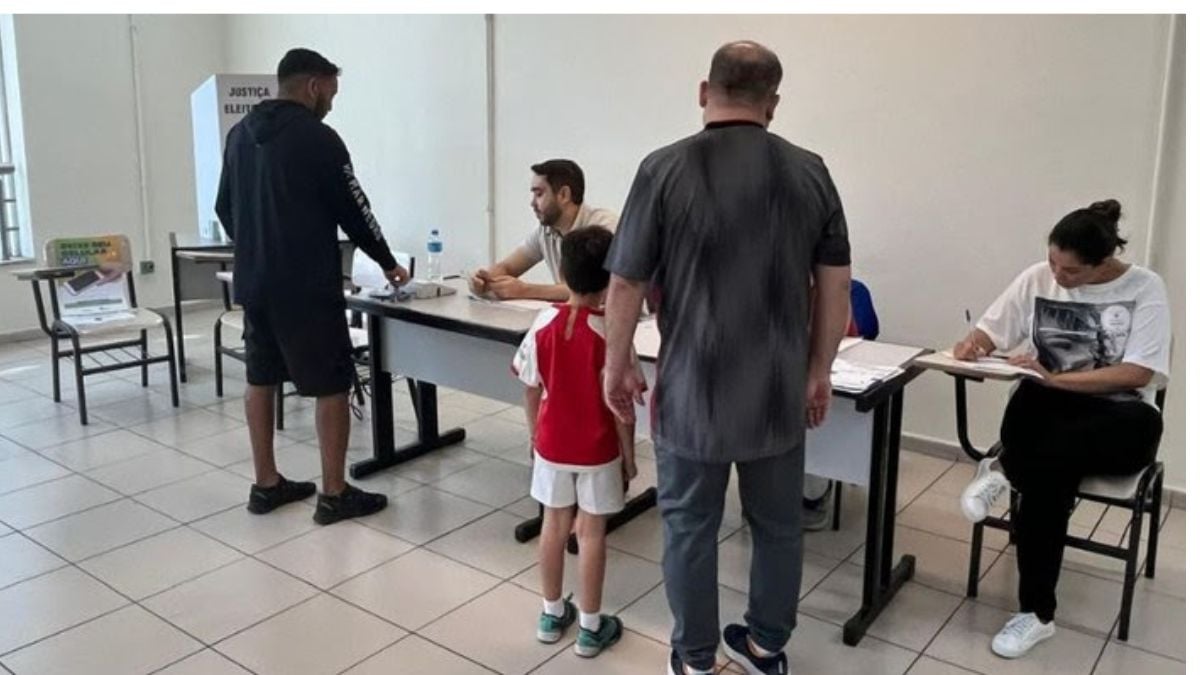 Pessoas em uma sala de votação, com agentes e eleitores sentados e em pé diante de mesas e urnas, tema relacionado a mesário 2026 e convocação para atuar nas eleições.