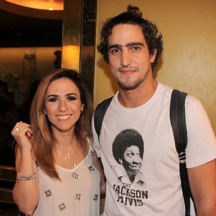 Tatá Werneck e Renato Góes aparecem sorrindo em foto de evento, ao lado um do outro, com Tatá ao fundo à esquerda e Renato à direita, usando camiseta com estampa de retrato.