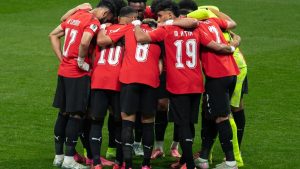 Torcedores egípcios se abraçam em campo após a partida, enquanto jogadores vestem uniformes vermelhos e celebram em meio à comemoração.
