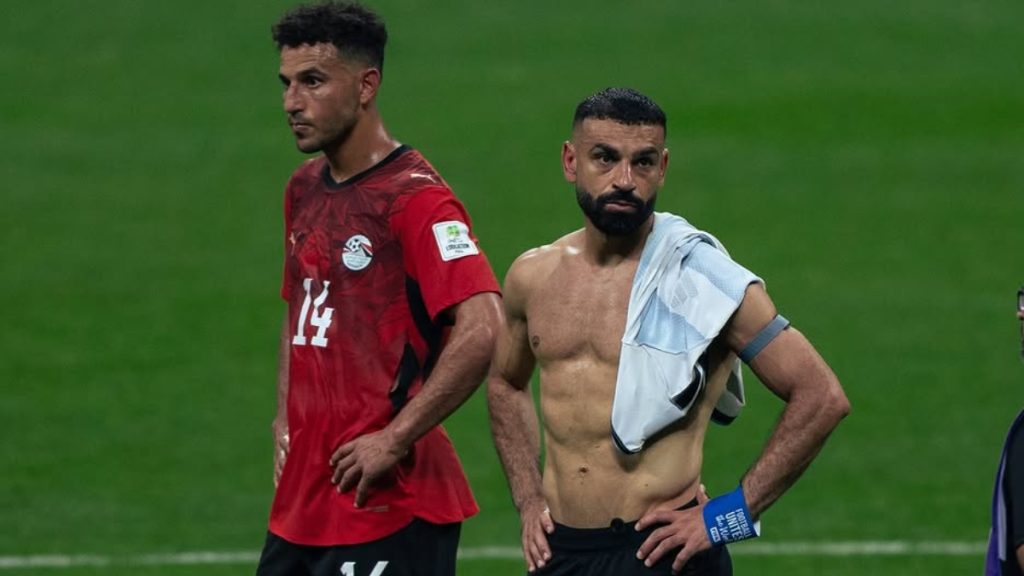 Dois jogadores durante partida em campo verde: um atacante da seleção do Egito com camisa vermelha e, ao lado, outro atleta sem camisa com toalha no ombro, em momento de pressão no jogo.