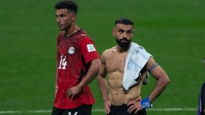 Egito em campo durante partida de futebol: jogador com camisa vermelha e outro atleta com toalha branca e braçadeira, em meio a gestos e tensão no gramado
