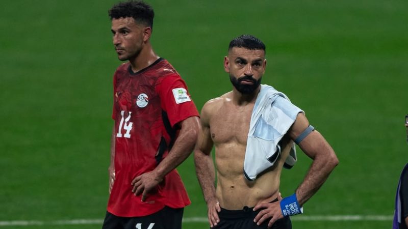 Egito em campo durante partida de futebol: jogador com camisa vermelha e outro atleta com toalha branca e braçadeira, em meio a gestos e tensão no gramado