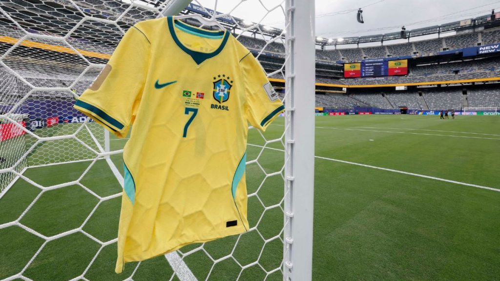 Camiseta amarela da Seleção Brasileira com número 7 exposta atrás das redes de um estádio, com o gramado ao fundo, em foto da CBF. Texto em destaque sugere discussão sobre dinheiro da Copa do Mundo.