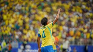Jogador brasileiro em campo levanta o braço comemorando durante uma partida da seleção, com arquibancada lotada em amarelo e verde ao fundo. Contexto sobre o prêmio milionário e a pergunta “O Brasil ganhou dinheiro para perder a Copa do Mundo?”.