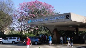 Feriado 9 de julho: fachada do Hospital de Clínicas com faixa de entrada e pessoas em frente ao prédio, em um dia ensolarado no campus.