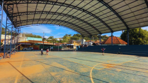 Vista interna de uma quadra esportiva coberta com estrutura metálica, piso quadriculado e linhas de marcação, em Águas de Lindóia, evidenciando que a reforma está abandonada e preocupa moradores.
