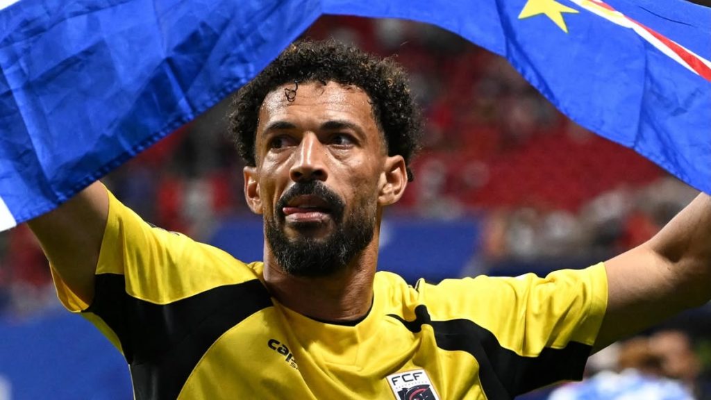 Vozinha em destaque: jogador de futebol celebra com os braços erguidos, usando uniforme amarelo com faixa preta, em partida com bandeira azul ao fundo.