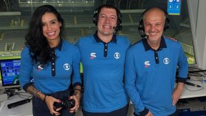 Equipe de locução em estúdio de transmissão, com três apresentadores usando headsets e camisas azuis, sorrindo diante de monitores na cobertura da Copa do Mundo da Globo.