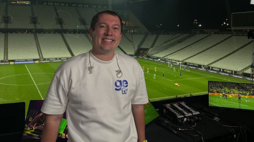 Imagem mostra narrador/locutor esportivo em pé no interior de um estádio de futebol, diante de monitores de transmissão, com camiseta do SporTV/GE TV, em contexto de Globo demite narrador.