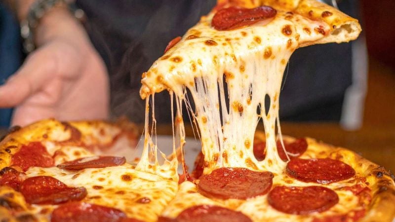Dia da pizza: close em uma pizza artesanal com fatias de pepperoni e queijo derretido, com uma porção sendo levantada e puxando fios de queijo em destaque.
