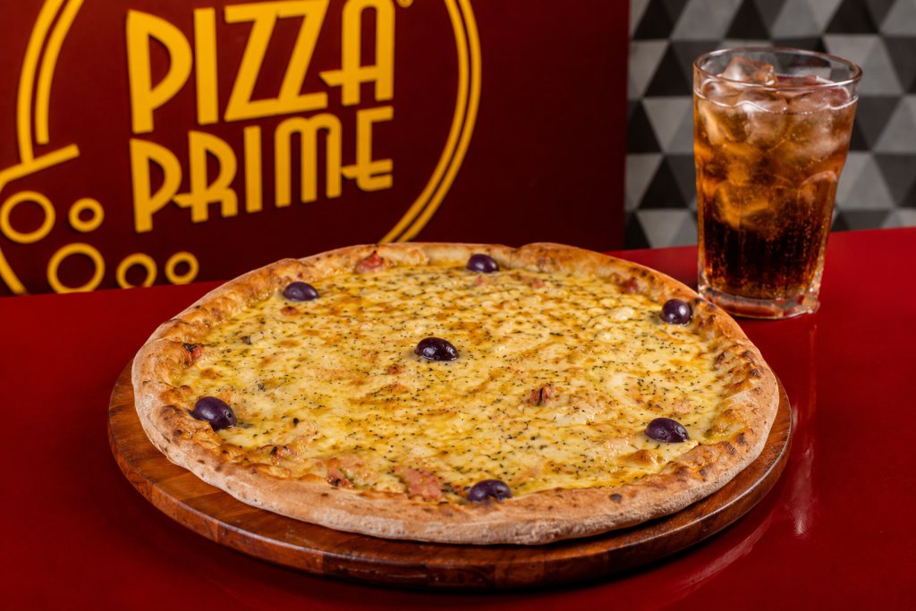 Dia da pizza: pizza tradicional tipo portuguesa em destaque sobre prato de madeira, com fundo promocional “Pizza Prime” e copo de bebida ao lado em ambiente de pizzaria.