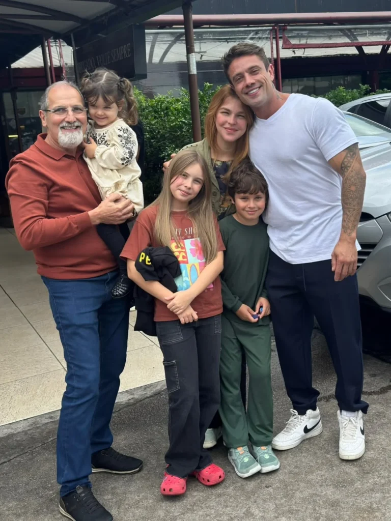 Rafael Cardoso posa em foto com a família em ambiente aberto, ao lado de crianças e de um idoso, sob cobertura próxima a carros.