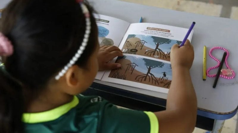 Criança participa de atividade de férias na biblioteca em Indaiatuba, com desenho em um livro aberto e lápis de cor durante programação gratuita de julho.
