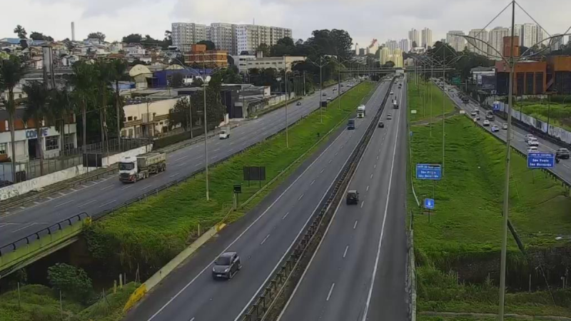Vista aérea de rodovia com faixas e canteiro central verde, carros e caminhões em circulação no Sistema Anchieta-Imigrantes durante obras, com sinalização e viadutos ao fundo, até 19 de julho.