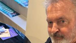Sam Neill aparece em close, com cabelos grisalhos e barba rala, olhando para a câmera em um ambiente interno. Imagem de perfil do ator conhecido por Alan Grant em Jurassic Park.