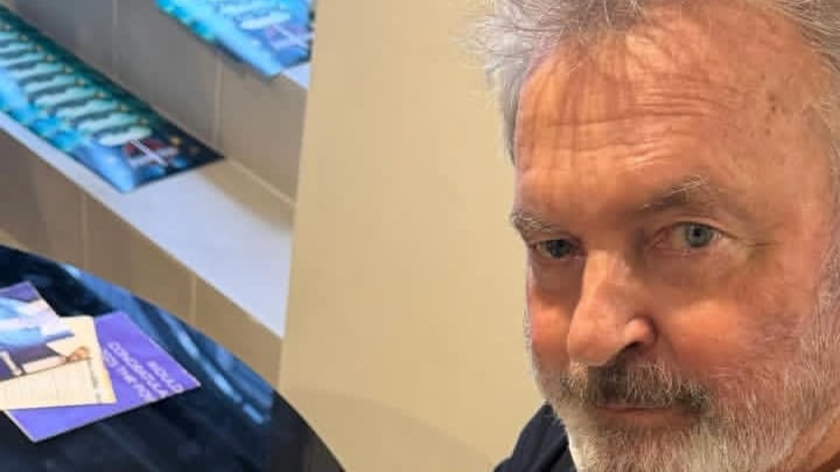 Sam Neill aparece em close, com cabelos grisalhos e barba rala, olhando para a câmera em um ambiente interno. Imagem de perfil do ator conhecido por Alan Grant em Jurassic Park.