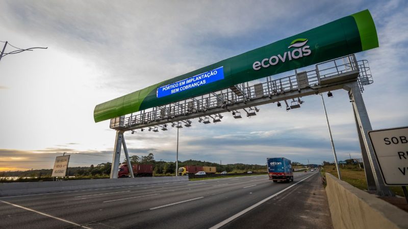 Pedágio eletrônico na Anchieta-Imigrantes com praça de cobrança automatizada e veículo passando sob pórtico verde da ecovias, ao entardecer.