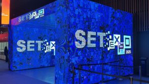Painel de LED azul com a marca “SET Expo 2026” em destaque, em um espaço de evento com estrutura iluminada, remetendo ao futuro da radiodifusão.