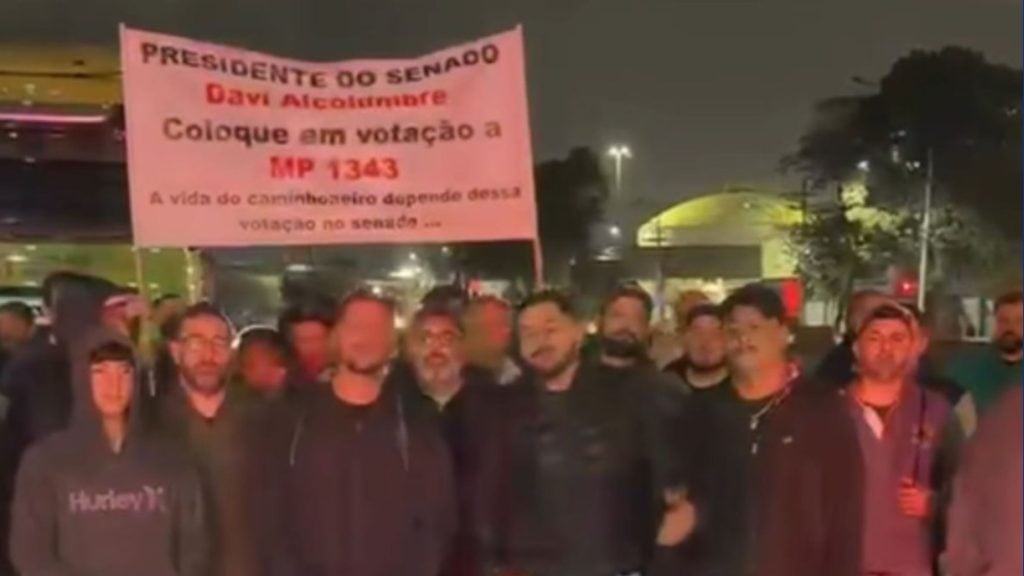 Greve caminhoneiros em frente ao Porto de Santos: grupo de manifestantes durante paralisação à noite, com cartaz vermelho ao fundo e iluminação do cais