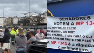 Caminhoneiros se reúnem em área portuária no litoral de SP durante Greve caminhoneiros, enquanto um caminhão com faixas e cartaz faz referência a mudanças no setor.