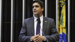 Cabo Daciolo em discurso na tribuna, usando terno escuro e gravata, ao lado de microfone, em ambiente institucional com bandeira ao fundo.