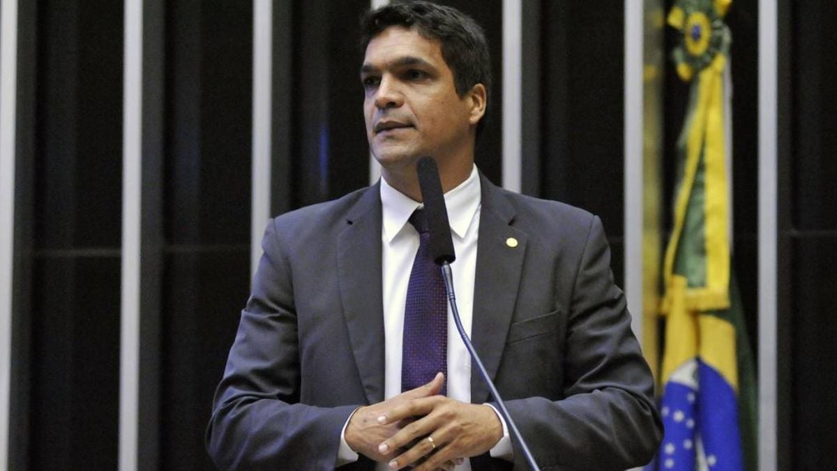 Cabo Daciolo em discurso na tribuna, usando terno escuro e gravata, ao lado de microfone, em ambiente institucional com bandeira ao fundo.