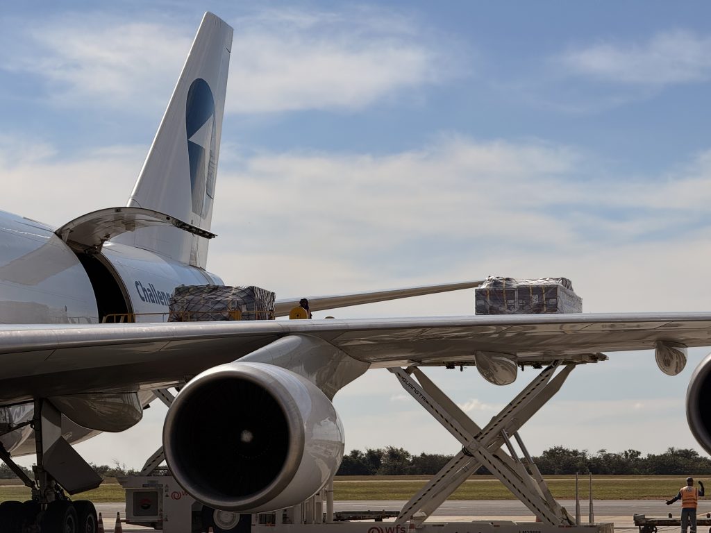 Close na lateral de um avião grande no aeroporto, com o motor e peças de carga visíveis sendo transportadas; referência à logística de shows do Harry Styles em SP e operação de transporte para o evento em Viracopos.
