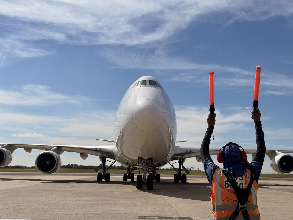 Harry Styles Viracopos: tripulante com sinalizadores vermelhos e um avião de grande porte na pista de aeroporto, em foto de logística para shows em São Paulo.