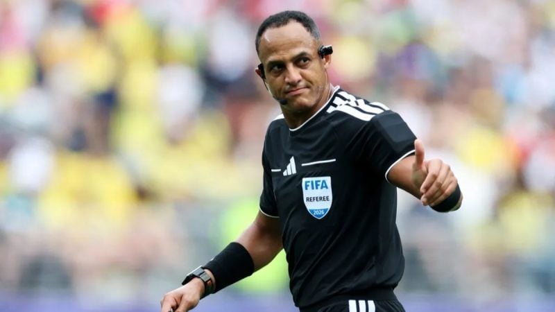 Ismail Elfath, árbitro FIFA, apita durante partida de futebol em um estádio, com gesto de mão em frente ao gramado e público ao fundo.