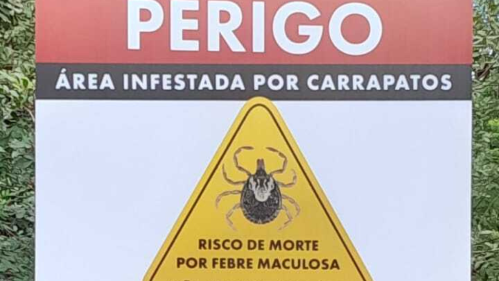 Placa de alerta “Perigo” com aviso “Área infestada por carrapatos” e “Risco de morte por febre maculosa”, indicando perigo associado à febre maculosa.