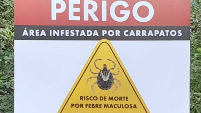 Placa de alerta “Perigo” com aviso “Área infestada por carrapatos” e “Risco de morte por febre maculosa”, indicando perigo associado à febre maculosa.