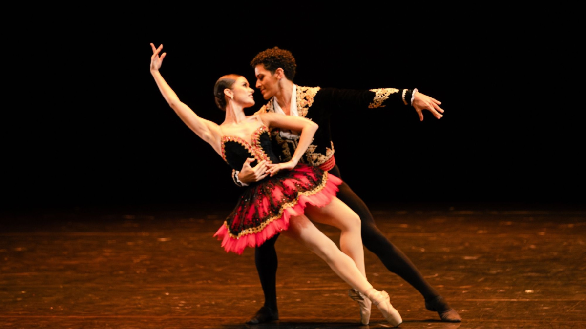 Dançarinos da São Paulo Companhia de Dança em cena: uma bailarina com saia colorida e tutu rosa e preto, apoiada em um parceiro, ambos com braços abertos, em palco iluminado, antes de uma apresentação gratuita em Santa Bárbara d’Oeste.