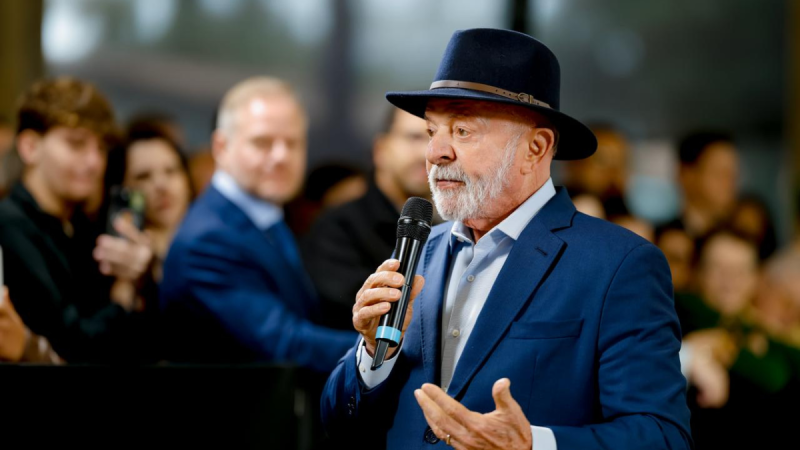 Lula discursa com microfone em evento, usando chapéu e terno azul, e critica jogadores da Seleção Brasileira ao comentar desempenho no futebol: “Se tivesse ganho, estava todo mundo dançando”.