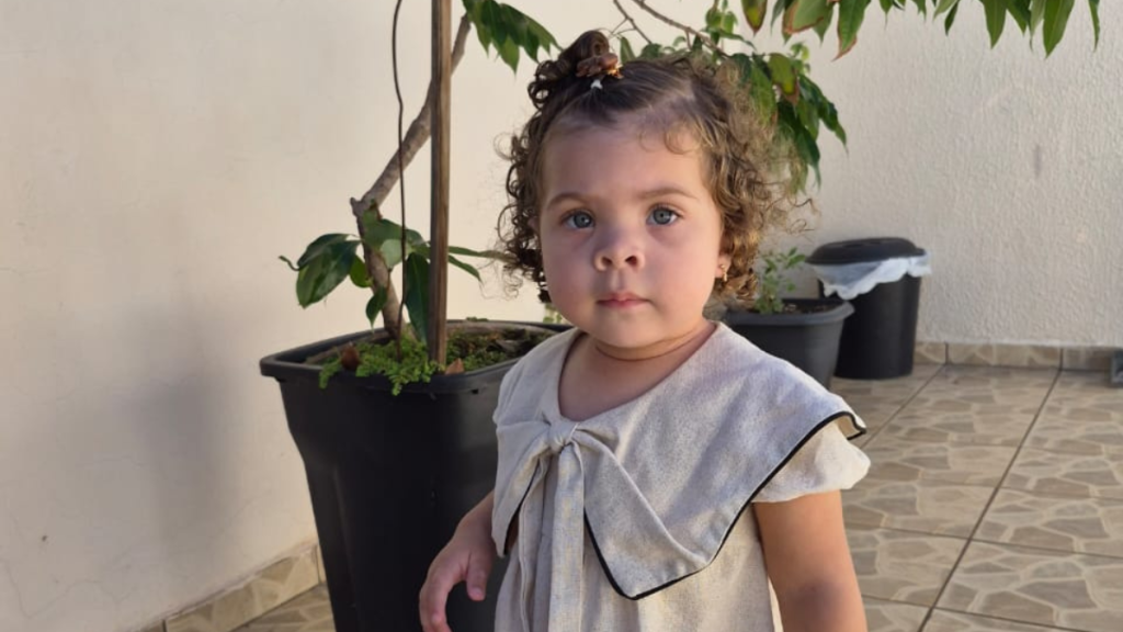 Heloise, uma menina de 1 ano e 10 meses, posa em ambiente externo ao lado de plantas em vasos; a imagem destaca o suporte do uso de tri-heptanoína para VLCAD
