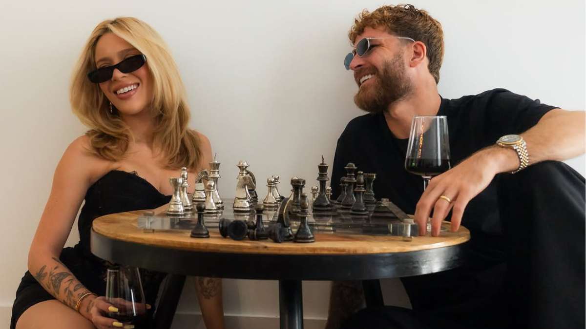 Karoline Lima e Léo Pereira em clima romântico, sentados lado a lado em um encontro com mesa de xadrez e taças de vinho, sorrindo como em confirmação de noivado e reconciliação.